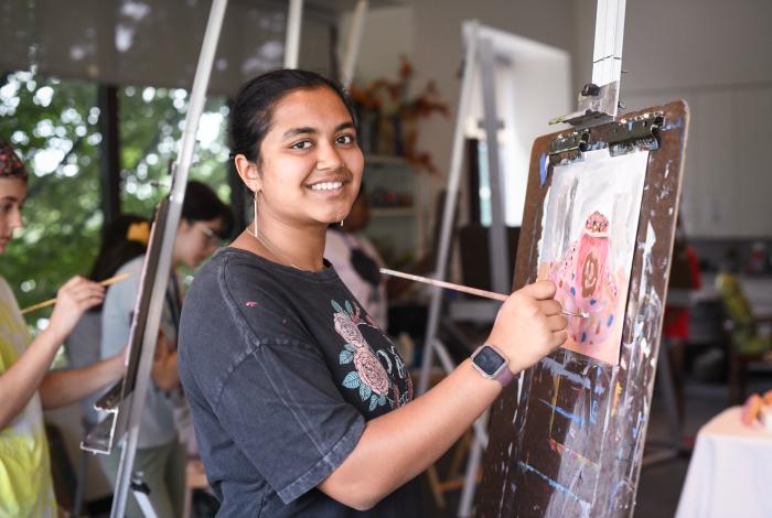 A teen student smiling at the camera as they paint on an easel